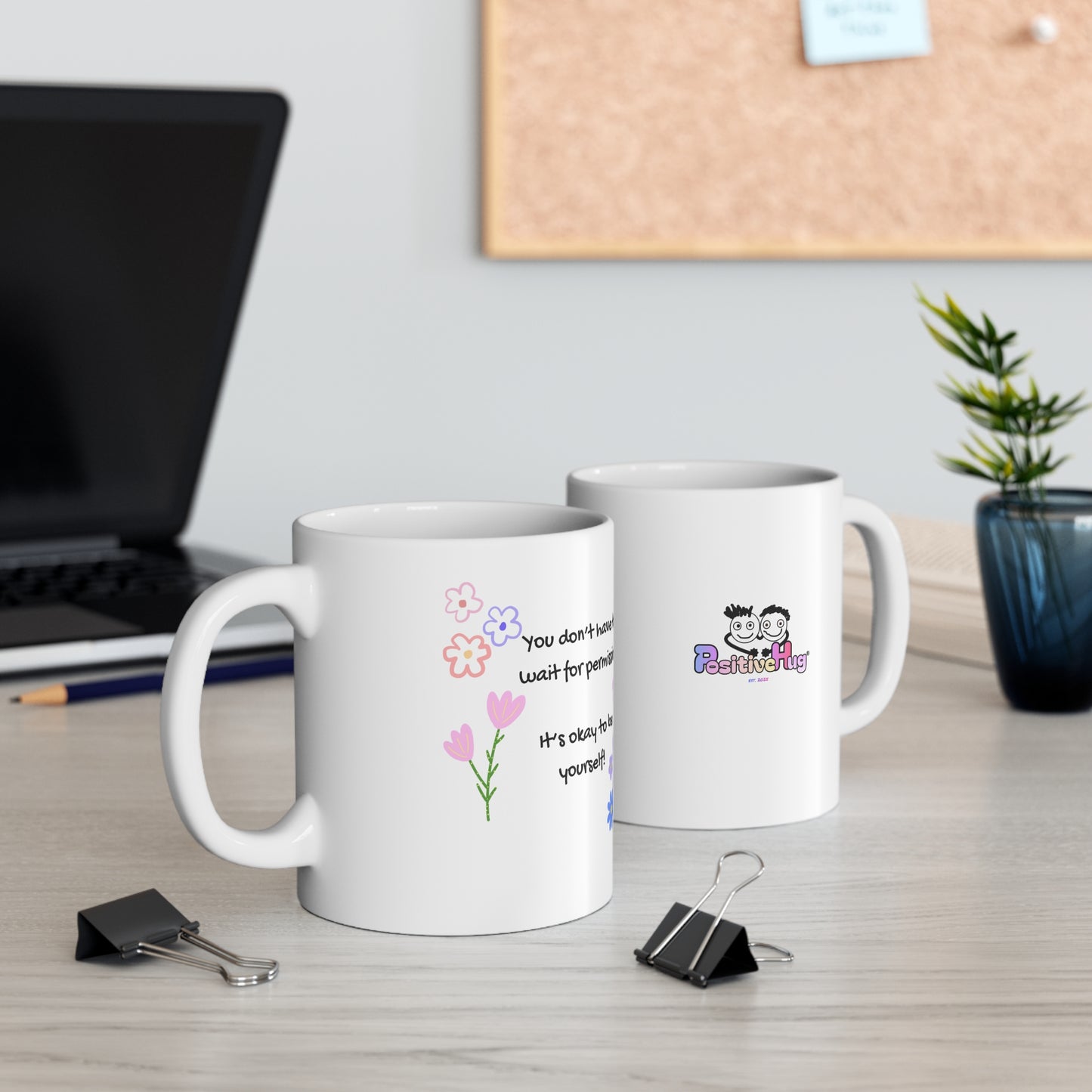 Two white mugs with printed designs on a desk with office supplies and a plant.