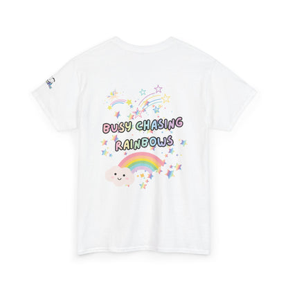 A white short-sleeve t-shirt with a colorful graphic on the front featuring the text 'BUSY CHASING RAINBOWS' in pastel bubble letters, surrounded by a smiling cartoon cloud, a rainbow, and stars.