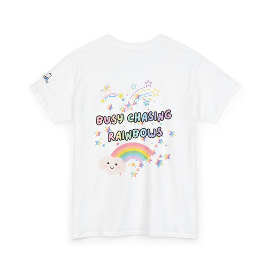 A white short-sleeve t-shirt with a colorful graphic on the front featuring the text 'BUSY CHASING RAINBOWS' in pastel bubble letters, surrounded by a smiling cartoon cloud, a rainbow, and stars.