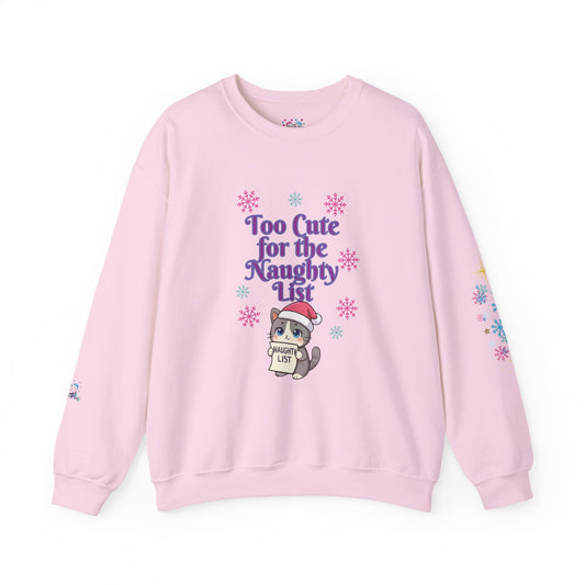 A light pink crewneck sweatshirt featuring a cute cartoon cat wearing a Santa hat and holding a sign that reads 'Cute List' surrounded by purple text that says 'Too Cute for the Naughty List' and colorful snowflake graphics.